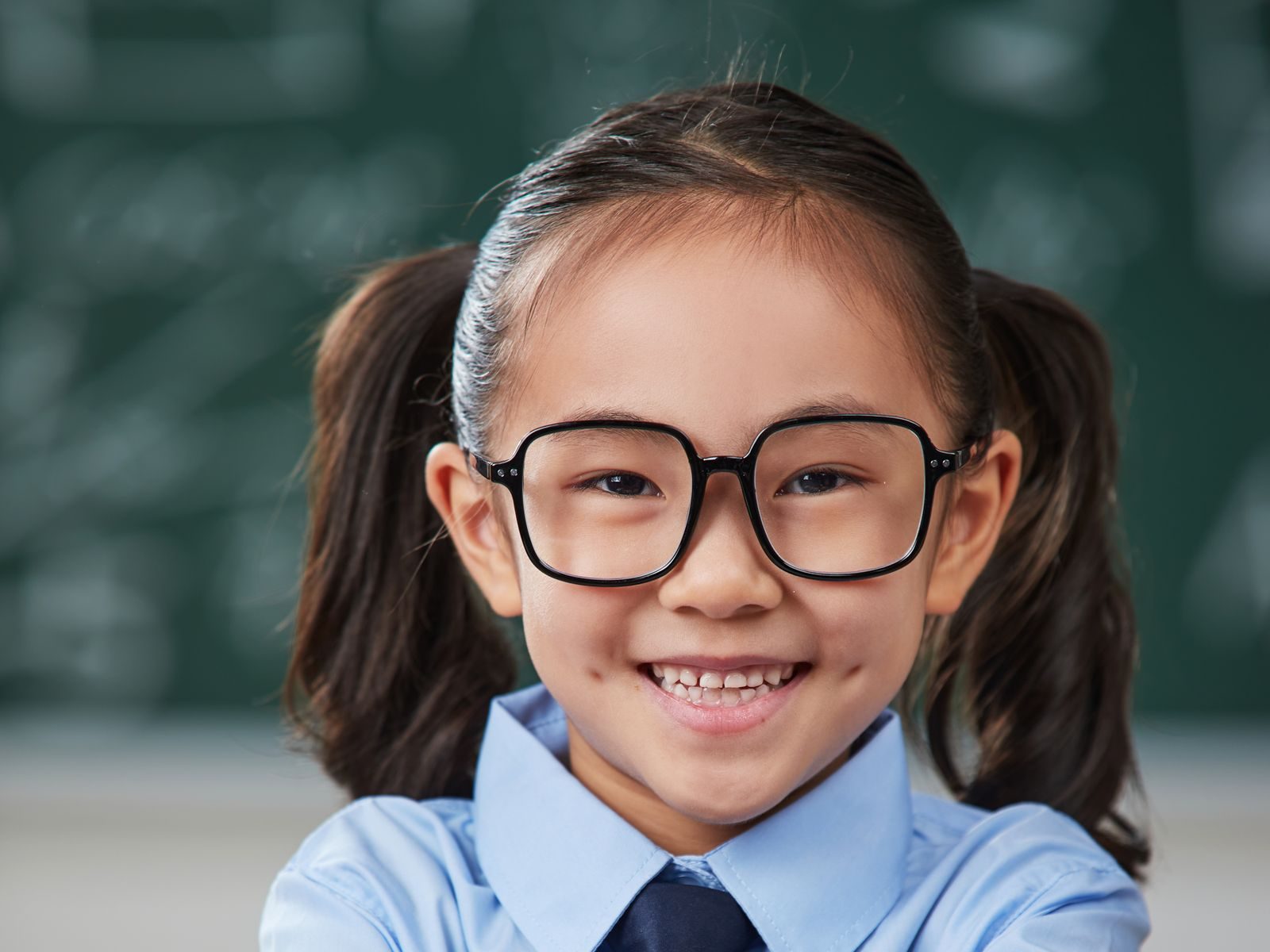 A smiling schoolgirl in glasses sitting at her desk in front of a chalkboard